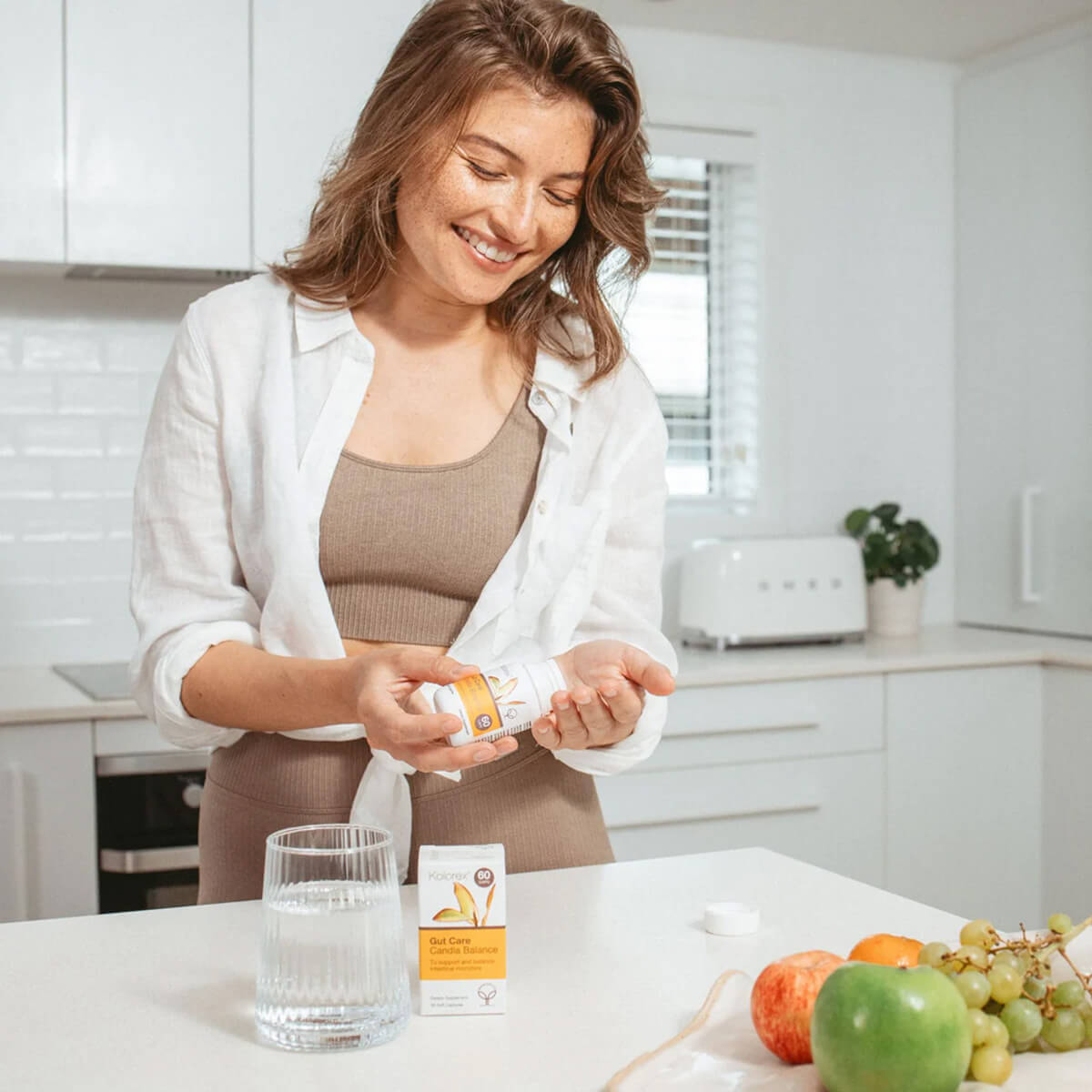 Woman in a kitchen holding a supplement bottle with a glass of water and fruits on the counter.