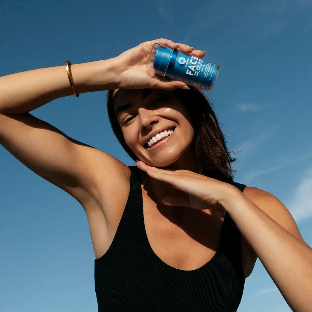 A woman outdoors under a clear blue sky holds a blue bottle of sunscreen above her head like a shield. She is wearing a black ribbed tank top and a gold bracelet.