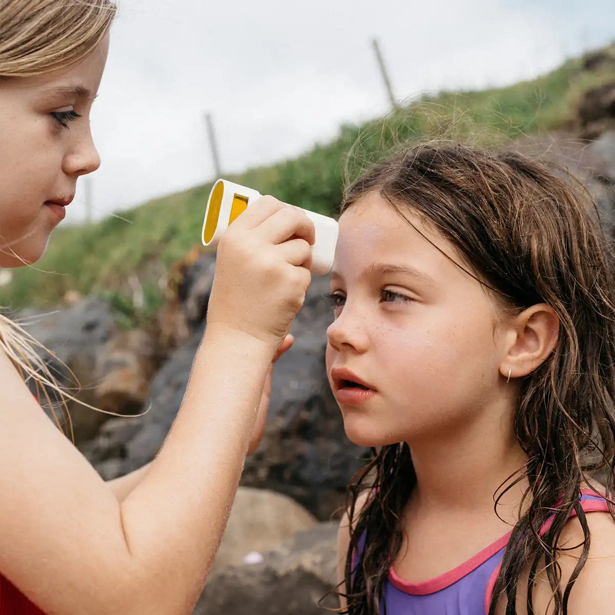 Two young girls outdoors, one applying sunscreen to the other's face.
