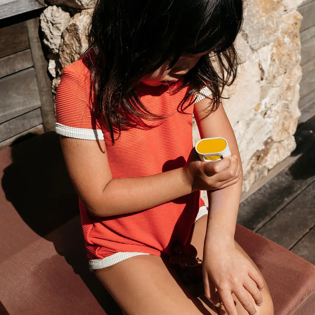 A child wearing a bright coral-orange ribbed short-sleeve romper with white trim sits outdoors on a cushioned bench, applying yellow sunscreen to their arm.