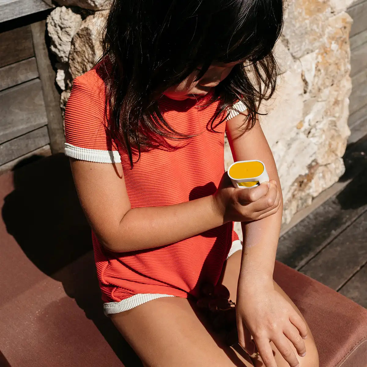 A child wearing a bright coral-orange ribbed short-sleeve romper with white trim sits outdoors on a cushioned bench, applying yellow sunscreen to their arm.