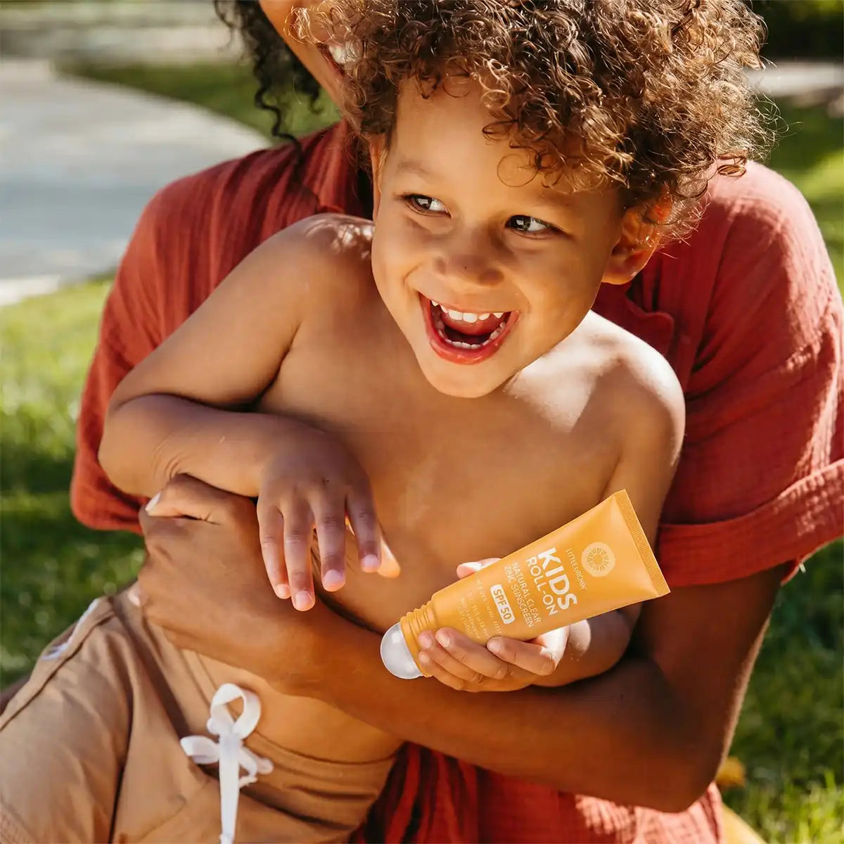 A smiling young child with curly hair is held outdoors by an adult wearing a rust-colored shirt. The child is holding a tube of kids sunscreen in a warm yellow-orange shade.