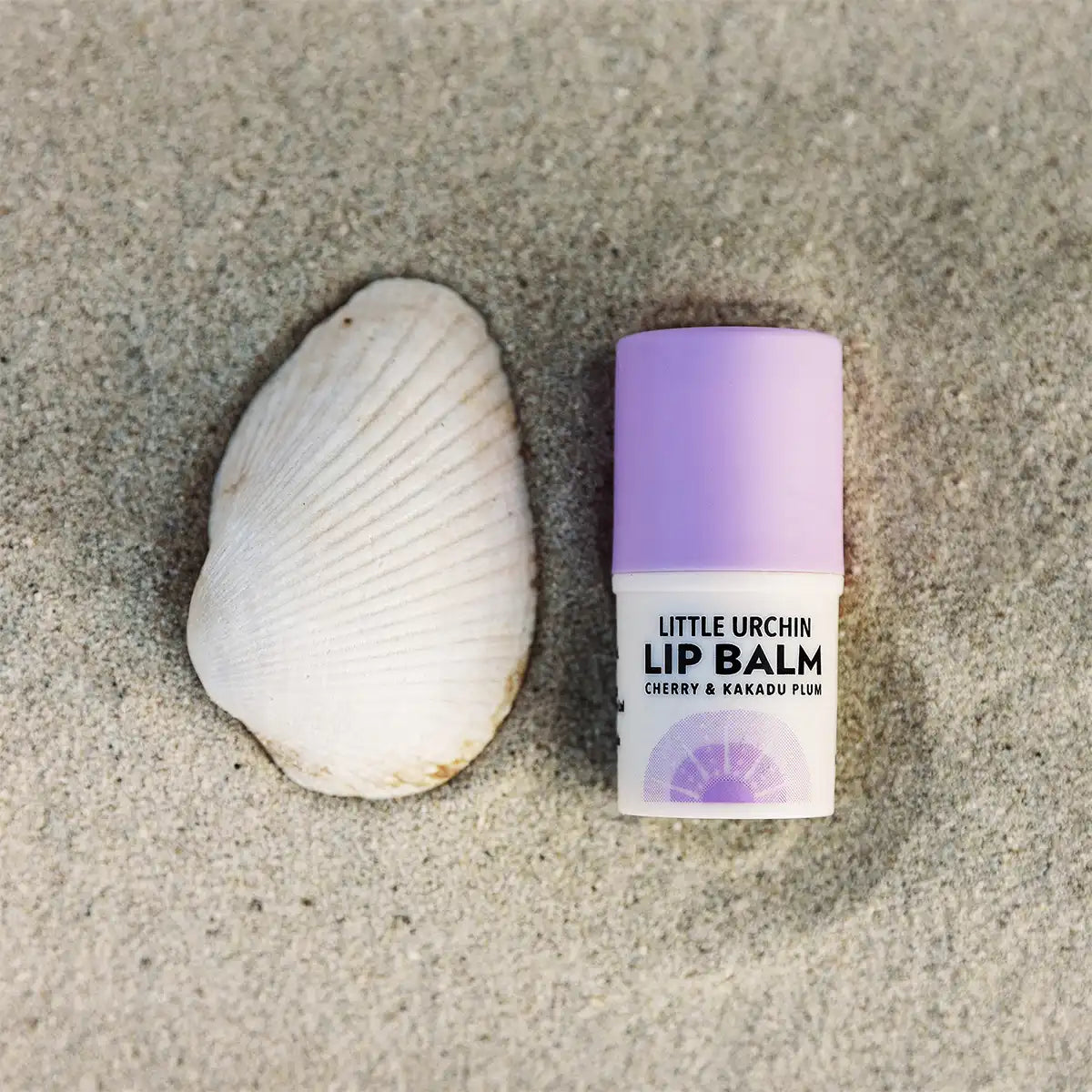 A small tube of lip balm with a purple cap on sand next to a white clam shell.