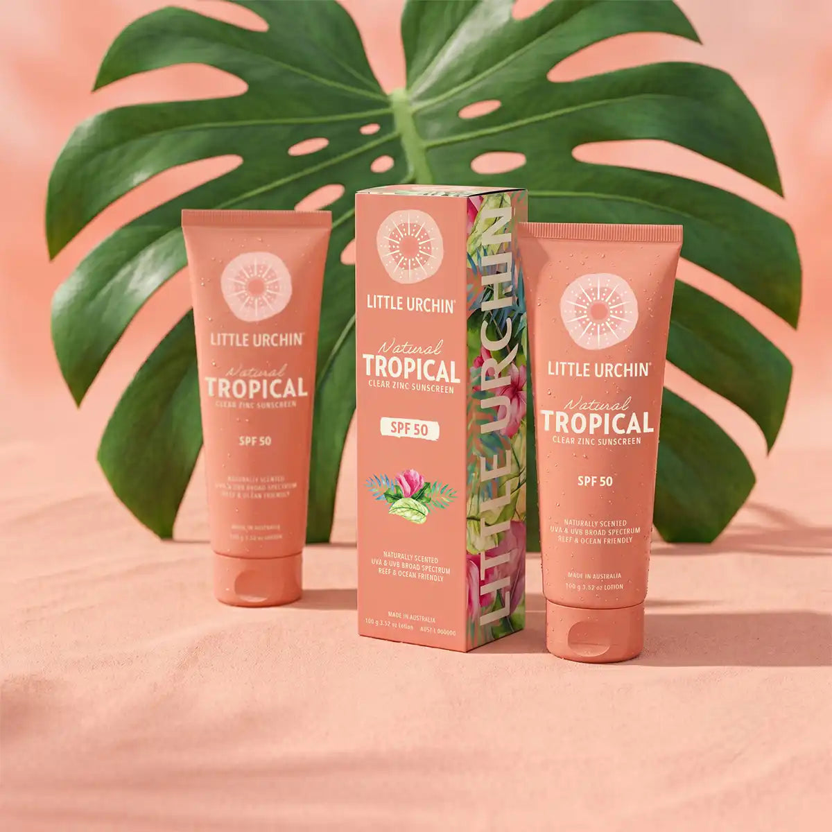 Little Urchin Natural Tropical Sunscreen Lotion SPF 50 in a peach squeeze tube, shown alongside its matching floral-patterned box and a second tube, styled with a large green monstera leaf on a peach fabric background.