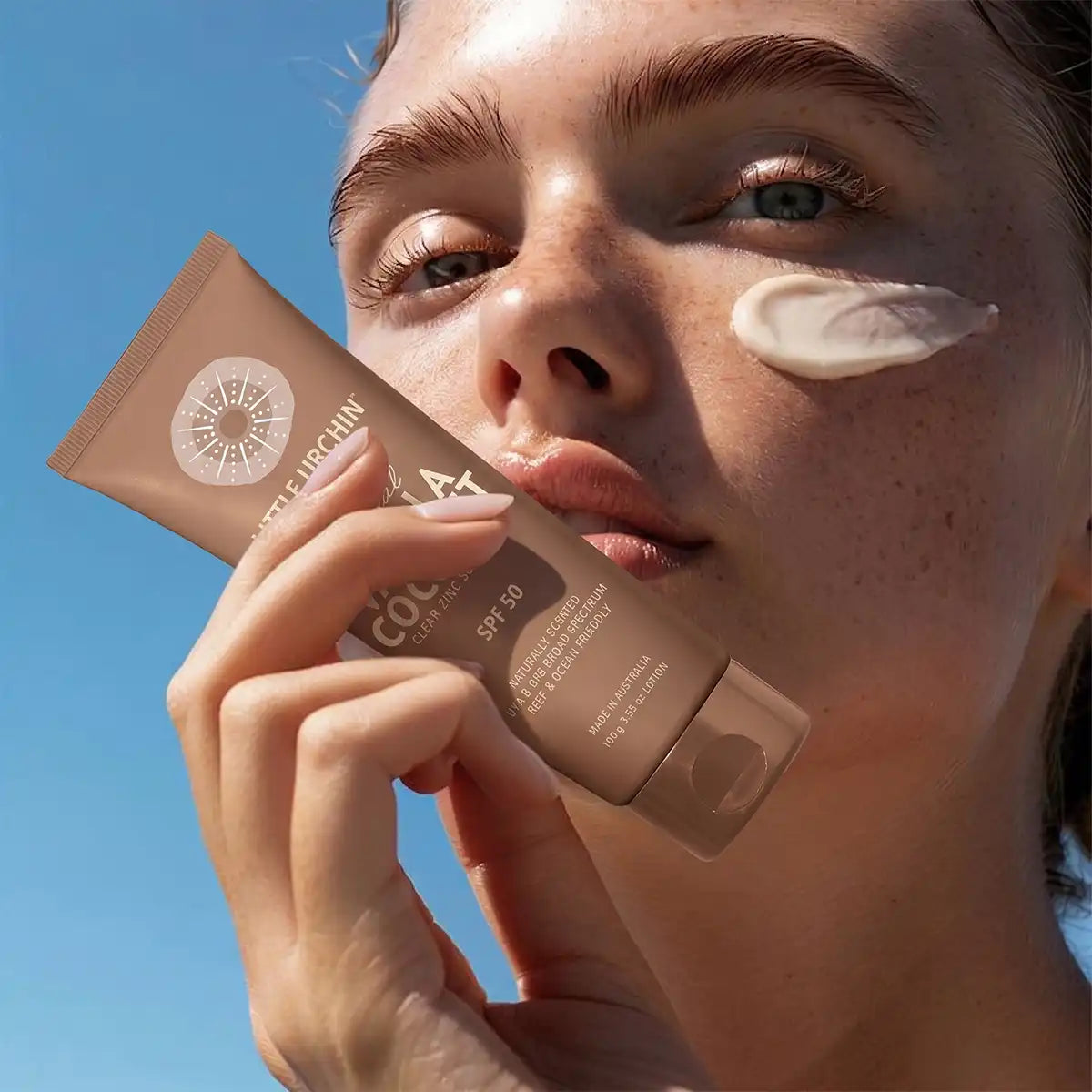 A close-up lifestyle shot of a woman outdoors under a clear blue sky, holding a tube of sunscreen next to a smear of cream on her cheek.