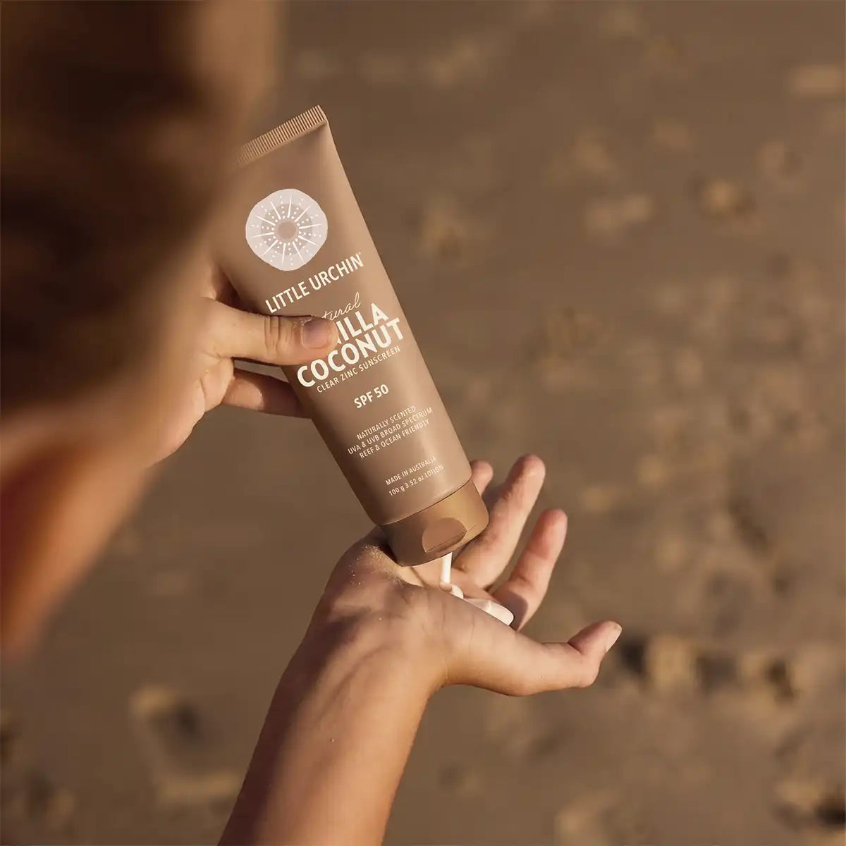 A person at a beach applies sunscreen from a brown tube onto their palm, with a warm sandy background.