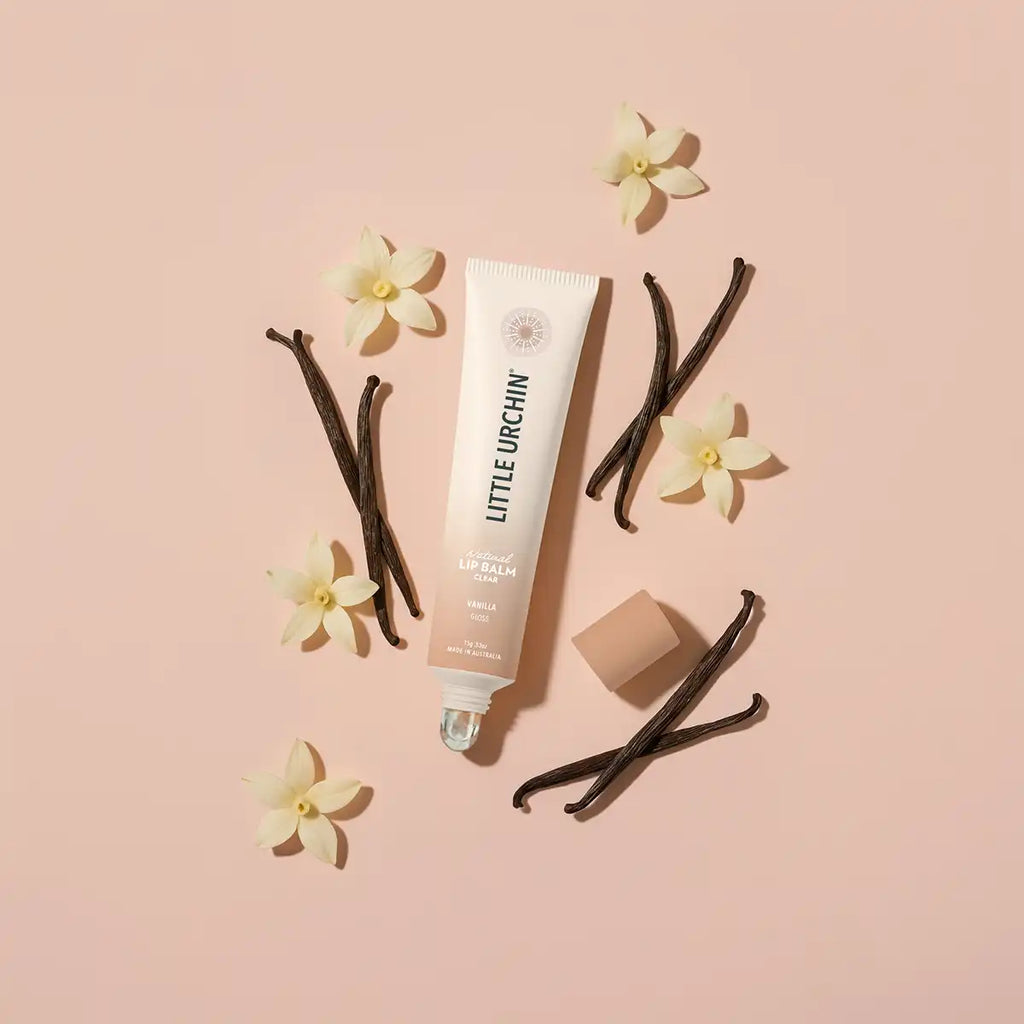 A tube of Little Urchin Vanilla lip balm is arranged on a soft pink background with vanilla beans, small white flowers, and the cap placed beside it.