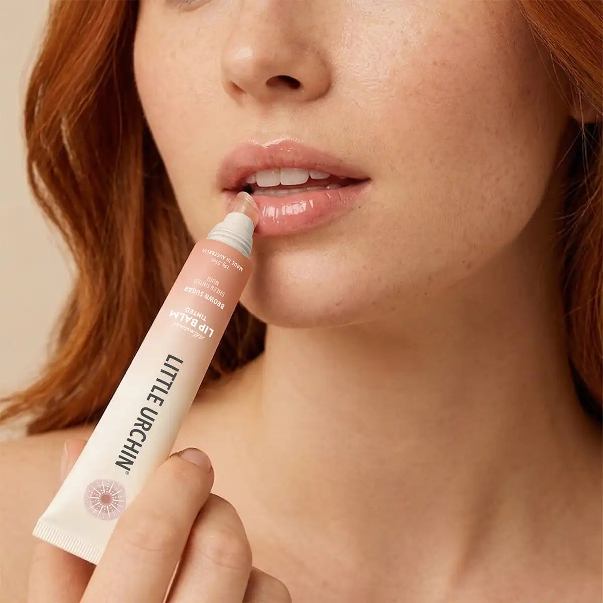 A close-up of a woman with red hair applying a peachy-pink lip balm from a Little Urchin tube to her lips.