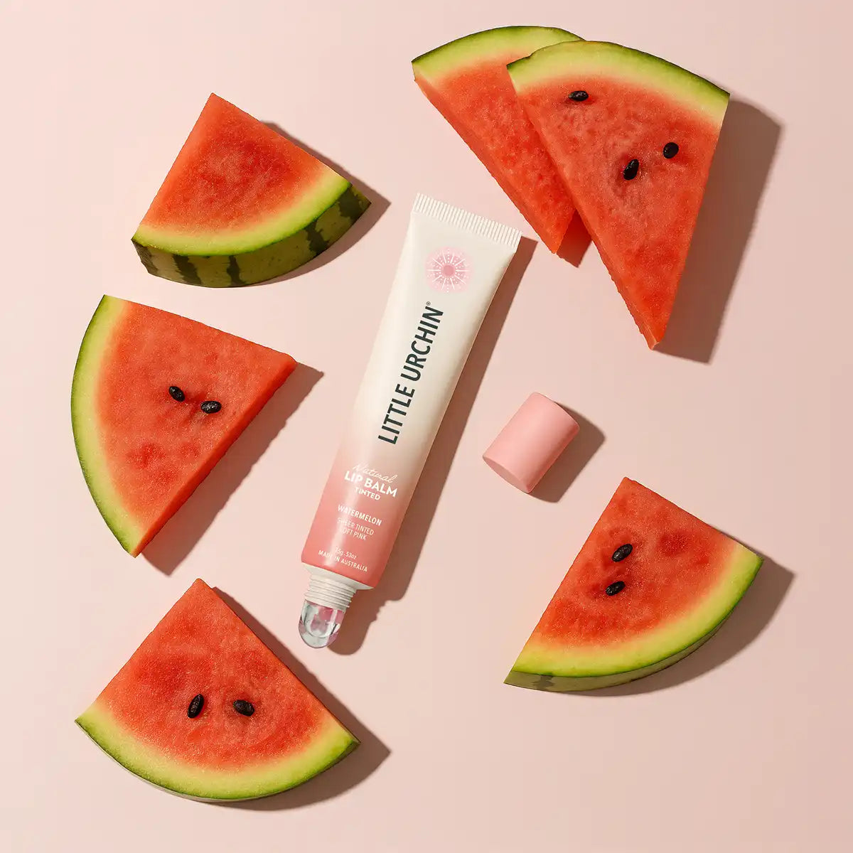 A tube of Little Urchin Lip Balm with a pink cap, styled in a flat-lay on a light pink background with watermelon slices arranged around it.