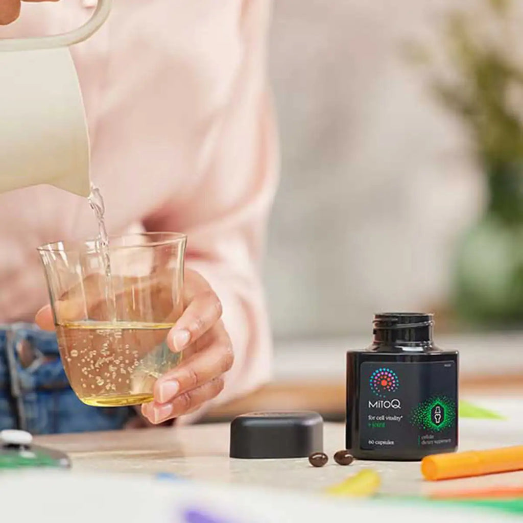 A person pouring hot water from a white pitcher into a glass beside a black supplement bottle.