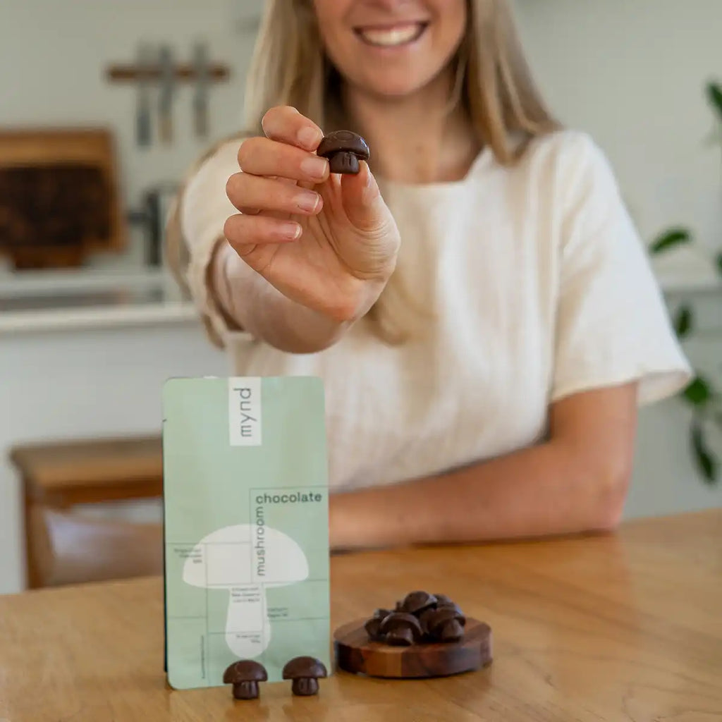 A woman in a white top holds up a small chocolate mushroom toward the camera, with a mint-green pouch several chocolate mushrooms on a wooden table in front of her.