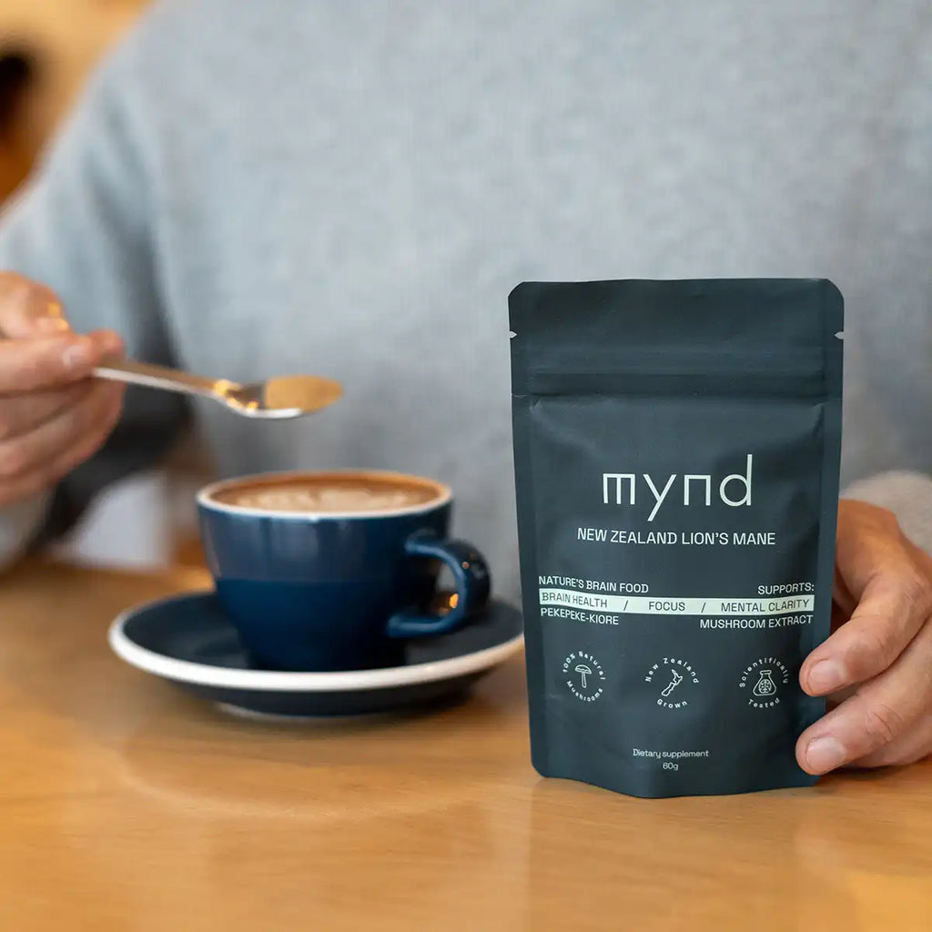 A person holds a spoon over a cup of coffee while holding a black stand-up pouch of Mynd New Zealand Lion's Mane supplement on a wooden table.