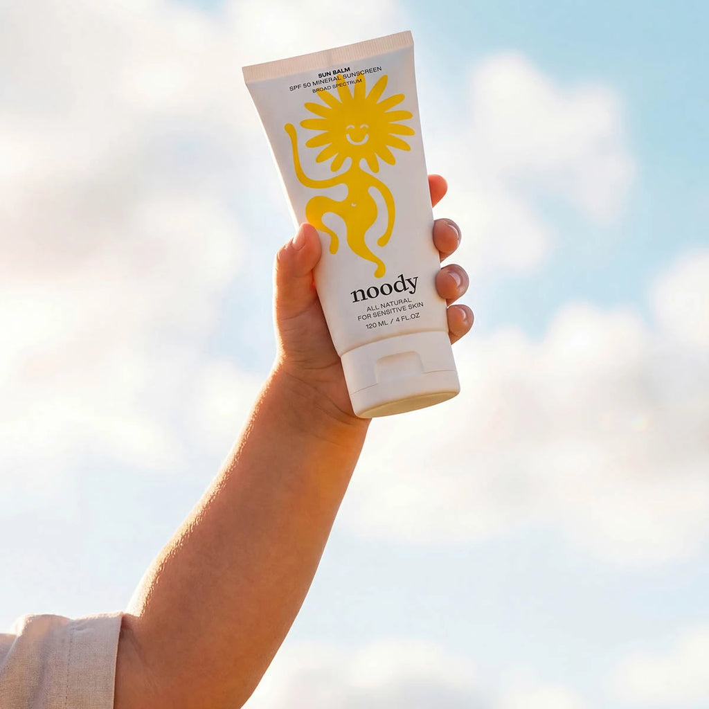 A hand holding a white tube of Noody Sun Balm sunscreen against a bright sky background.