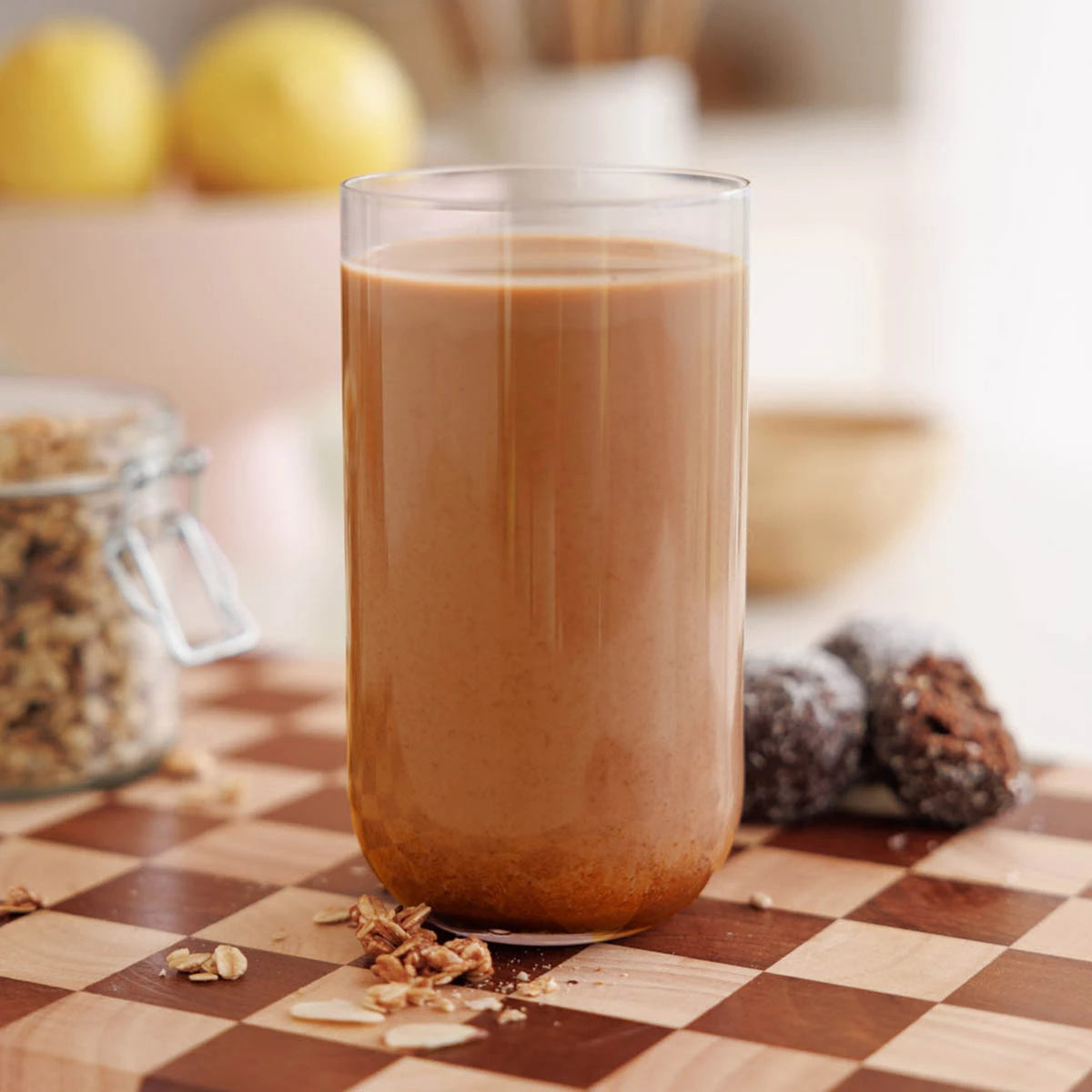 Glass of chocolate milk, made with plant protein powder, on a checkered tablecloth with granola and lemons in the background.