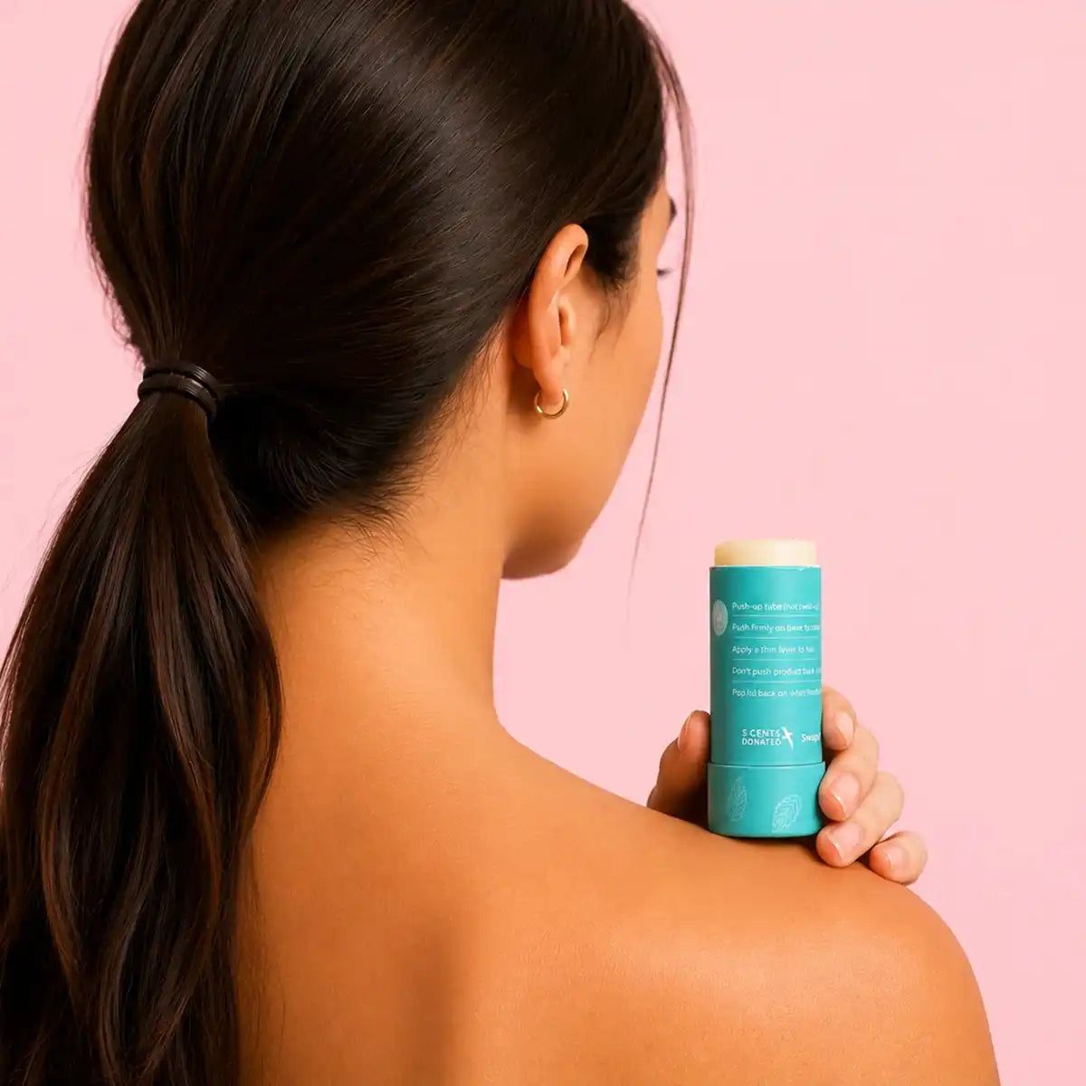 A woman with a dark ponytail, seen from behind, holds a teal slo hair wax stick against her bare shoulder on a pale pink background