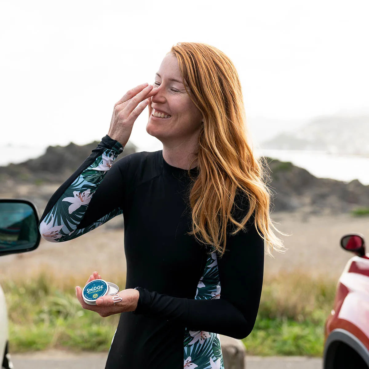 A woman outdoors near a beach or coastal road wearing a black long-sleeve wetsuit with green tropical leaf print on the sleeves and side panels. She is smiling and touching her face, holding a small round product container.