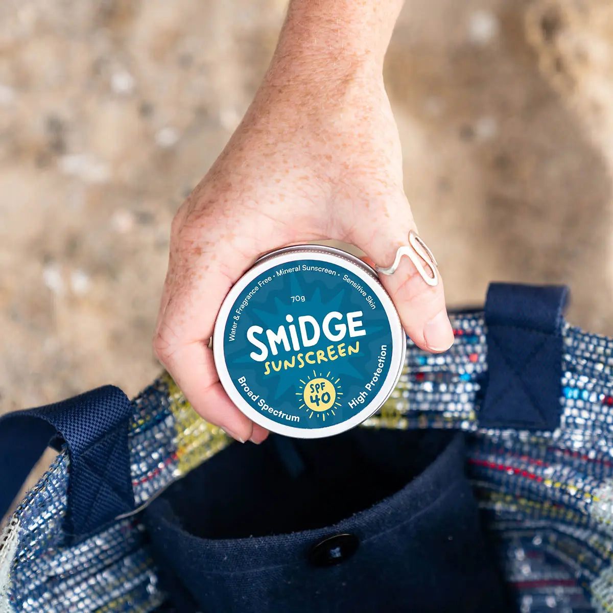 A hand holds a round tin of Smidge Sunscreen over a woven tote bag, with a sandy outdoor background.