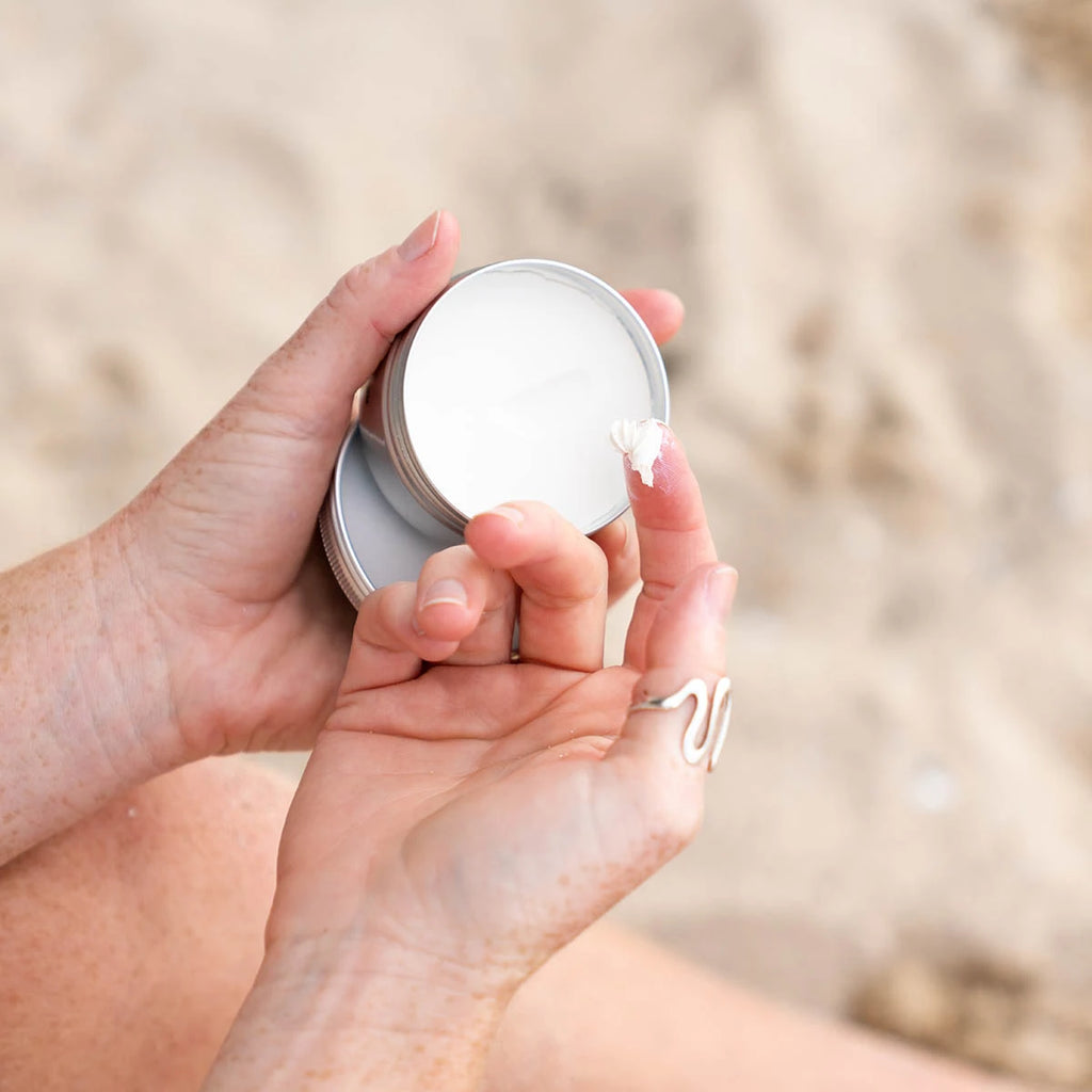 A close-up of a person's hands holding an open silver tin of white cream, with a small piece of shell on the lid, against a blurred sandy beach background.