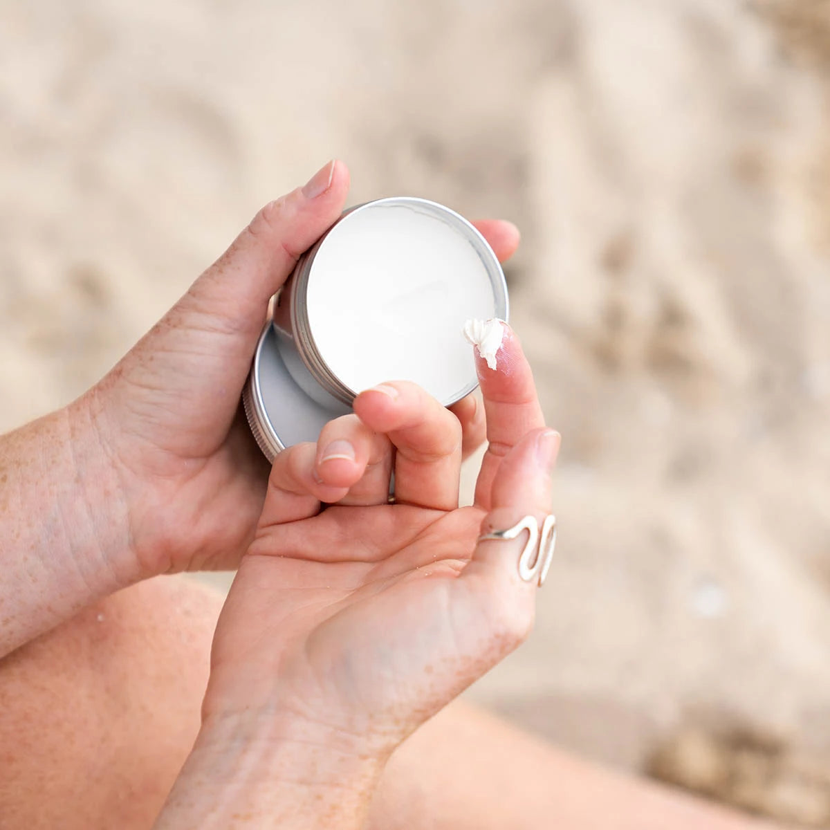 A close-up of a person's hands holding an open silver tin of white cream, with a small piece of shell on the lid, against a blurred sandy beach background.