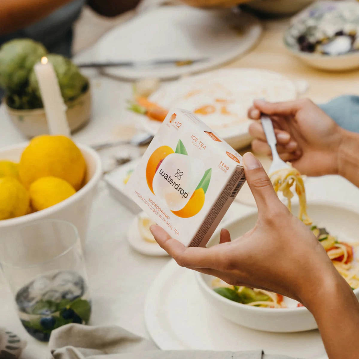 Person holding a box of Waterdrop tea at a dining table with food and drinks.