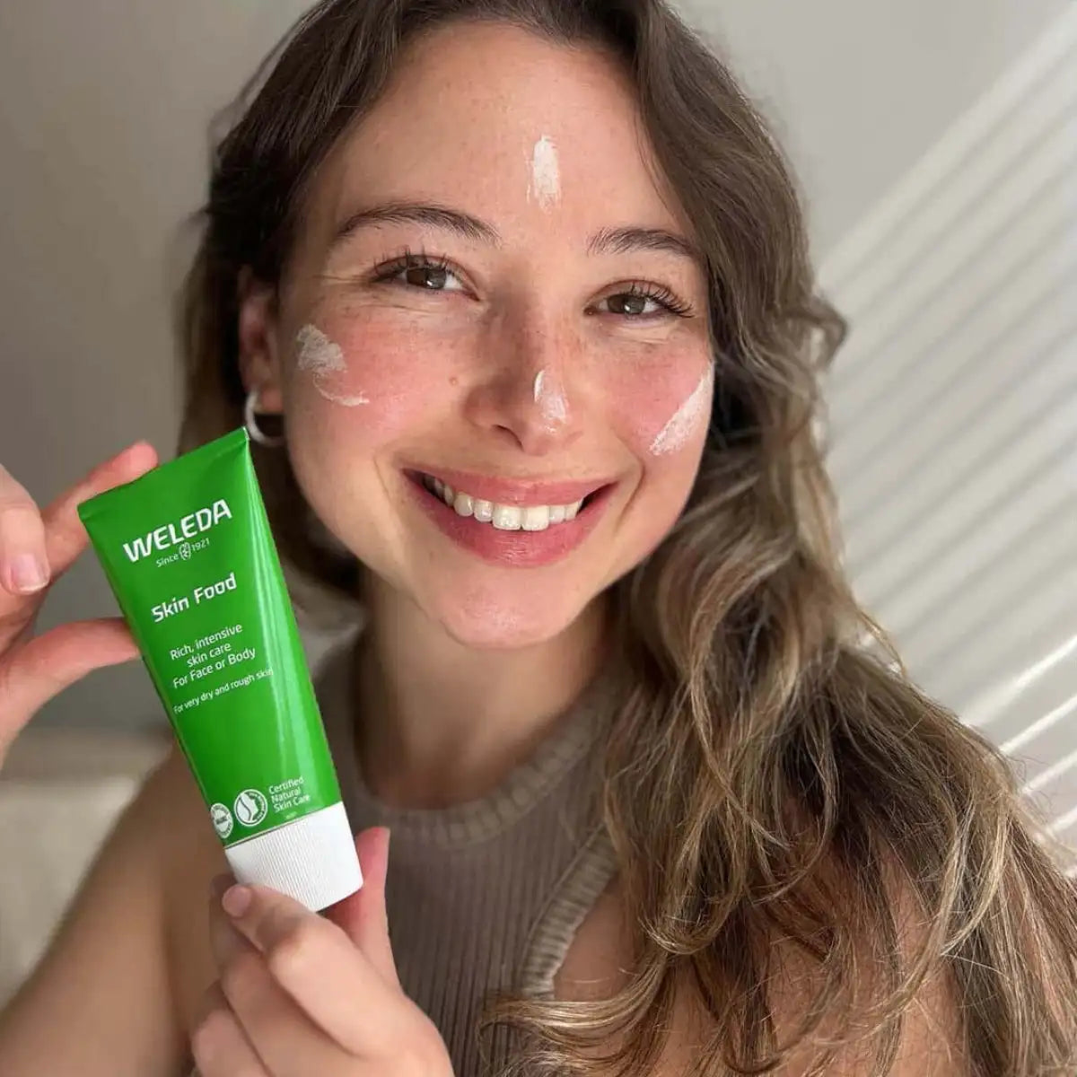 Woman holding up green tube of Weleda Skin Food with a few swatches on her face.