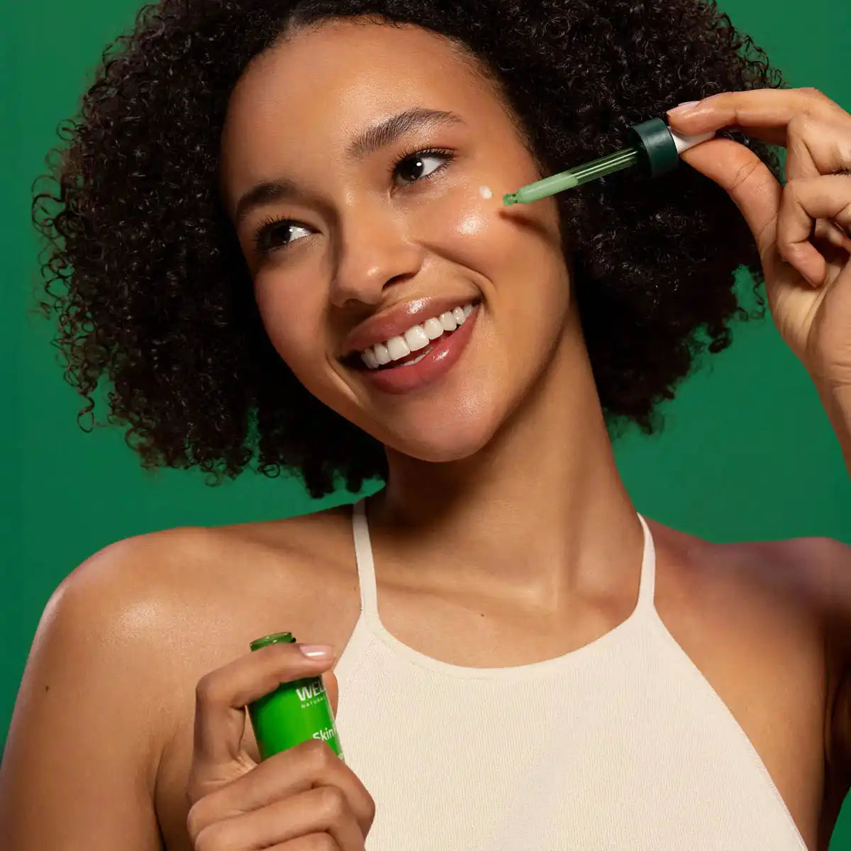 Woman applying Weleda Skin Food Super Serum to her cheek while smiling.  Green background that matches the product packaging.