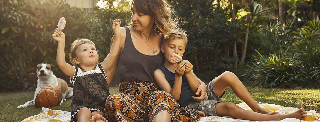 Mother Sits With Kids Outside On Picnic Blanket As They Eat Iceblocks