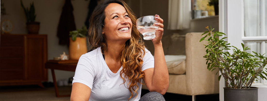 Woman smiles and holds glass of water as she sits outside 