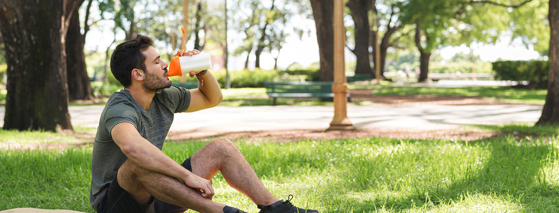 Man sits on grass in park as he drinks from orange protein shake bottle
