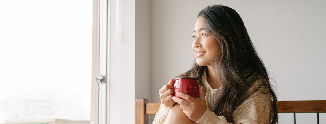 Woman holds beverage and smiles while looking out window 